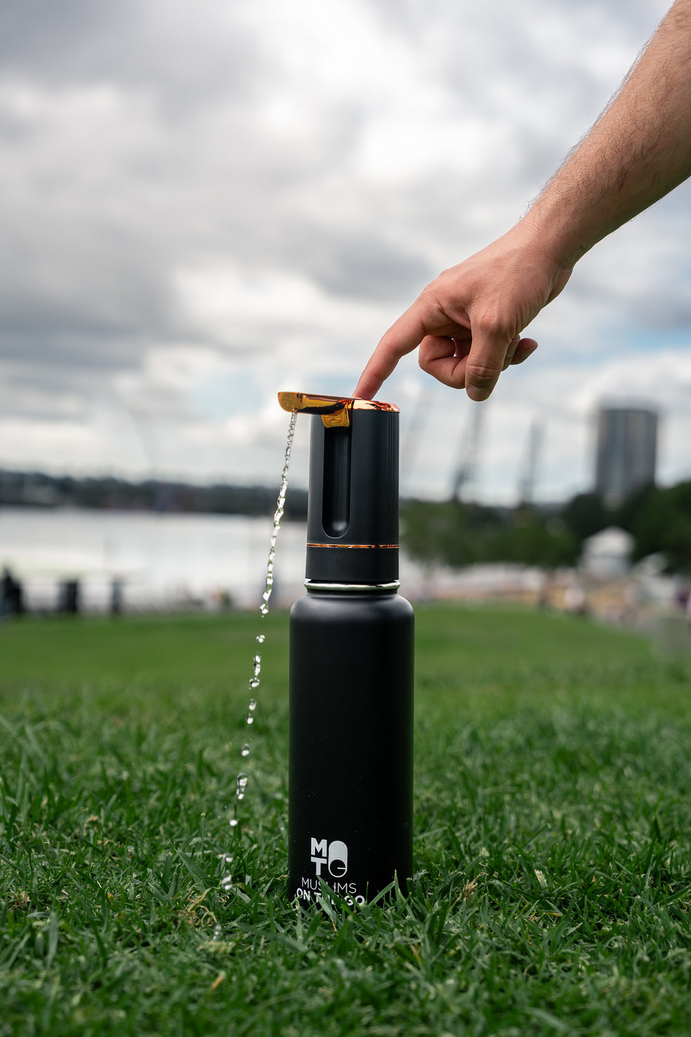 A black insulated bottle with a gold and black automatic water dispenser sits on green grass outdoors, with water flowing out. A person's hand is pressing a button to switch on the dispenser.