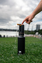 A black insulated bottle with a gold and black automatic water dispenser sits on green grass outdoors, with water flowing out. A person's hand is pressing a button to switch on the dispenser.